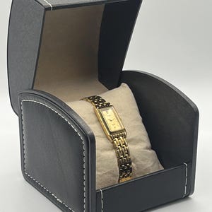 May include: A gold-toned wristwatch with a rectangular face and a linked band rests on a beige pillow inside a black watch box. The box is open, revealing the watch, and has white stitching around the edges.