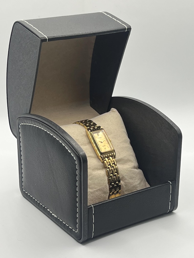 May include: A gold-toned wristwatch with a rectangular face and a linked band rests on a beige pillow inside a black watch box. The box is open, revealing the watch, and has white stitching around the edges.