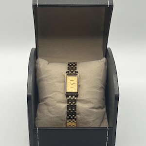 May include: A gold-toned rectangular watch with a matching band, presented in a black box with a light-colored interior. The watch features a gold face and is displayed on a beige pillow.