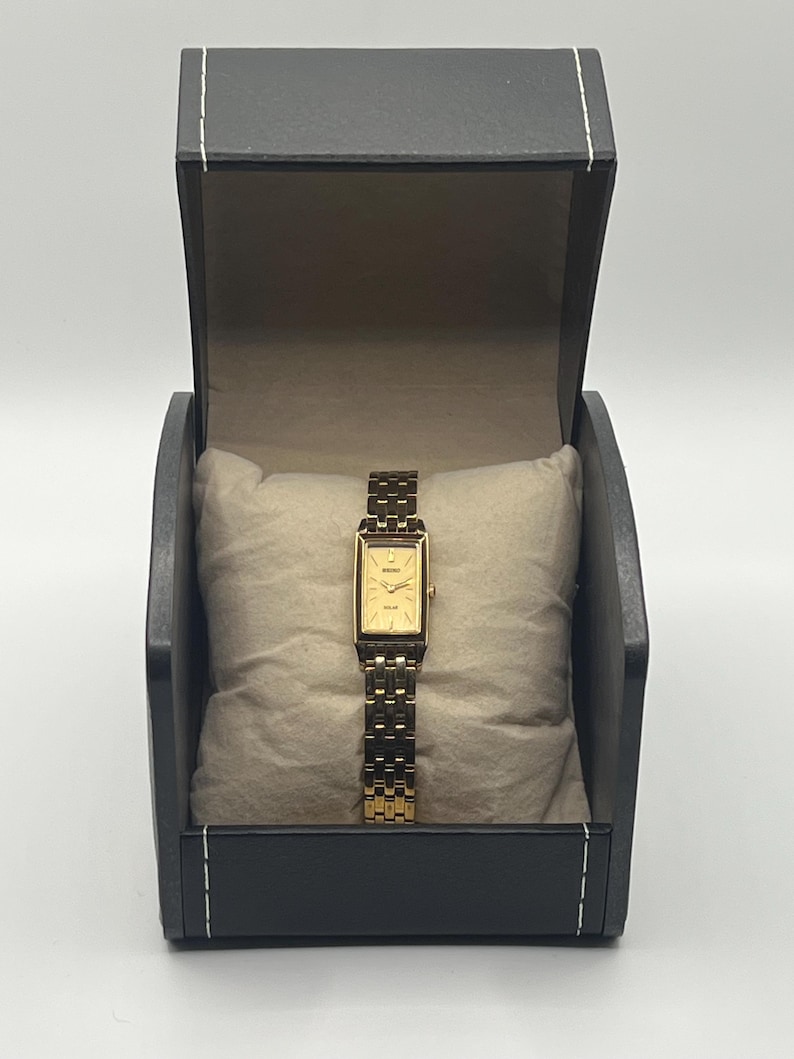 May include: A gold-toned rectangular watch with a matching band, presented in a black box with a light-colored interior. The watch features a gold face and is displayed on a beige pillow.