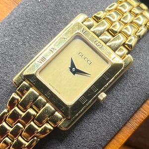 May include: A gold-toned Gucci watch with a rectangular face and a matching gold link bracelet. The watch face is a light gold color with black hands and Roman numeral hour markers. The Gucci logo is displayed on the watch face. "SWISS MADE" is printed at the bottom.