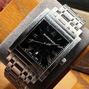 May include: A silver Burberry watch with a rectangular black face. The watch features Roman numeral hour markers and the Burberry logo. The watch band is made of silver links. The watch is displayed on a gray surface.