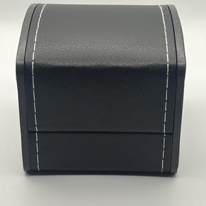 May include: A black, rectangular box with a curved top and a hinged lid. The box is made of a textured material and features white stitching along the edges. The box is likely designed for jewelry or other small items.