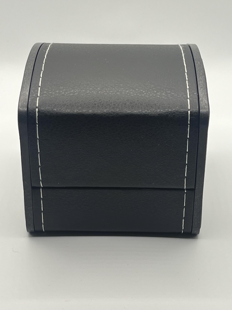 May include: A black, rectangular box with a curved top and a hinged lid. The box is made of a textured material and features white stitching along the edges. The box is likely designed for jewelry or other small items.
