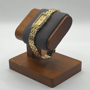 May include: A gold-toned rectangular wristwatch with a matching link bracelet is displayed on a wooden watch stand. The watch has a light-colored face with black hands and markers. The stand has a dark gray cushion.