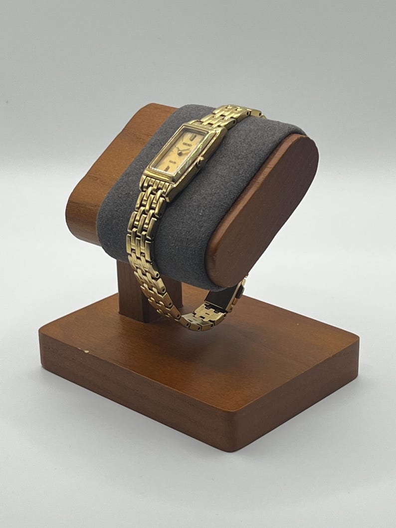 May include: A gold-toned rectangular wristwatch with a matching link bracelet is displayed on a wooden watch stand. The watch has a light-colored face with black hands and markers. The stand has a dark gray cushion.