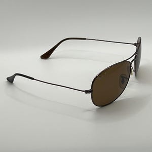 May include: Brown aviator sunglasses with a dark metal frame. The lenses are a warm brown color, and the temples curve gently. The Ray-Ban logo is visible on the lens. The sunglasses are set against a white background.