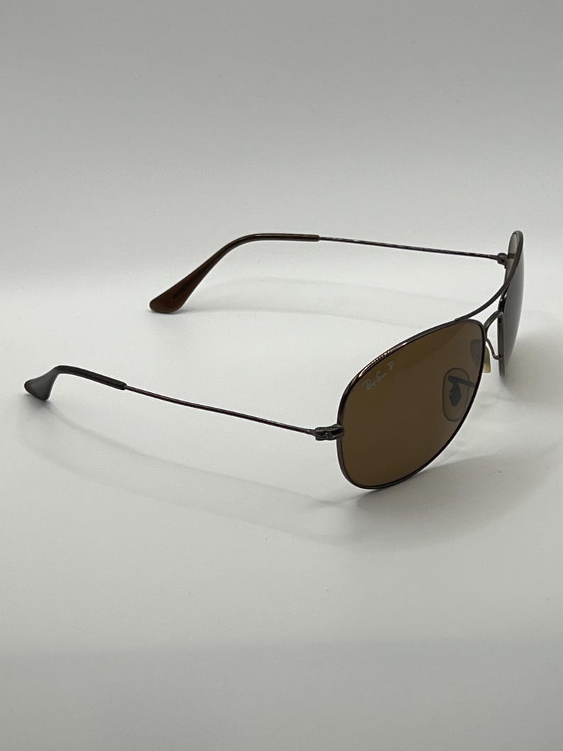 May include: Brown aviator sunglasses with a dark metal frame. The lenses are a warm brown color, and the temples curve gently. The Ray-Ban logo is visible on the lens. The sunglasses are set against a white background.