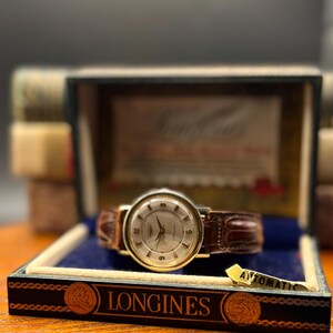 Puede incluir: Un reloj de pulsera autom&aacute;tico Longines vintage con una correa de cuero marr&oacute;n, presentado en su caja original. El reloj tiene una caja dorada y una esfera clara con marcadores de hora. La caja es negra con letras doradas y un interior azul.