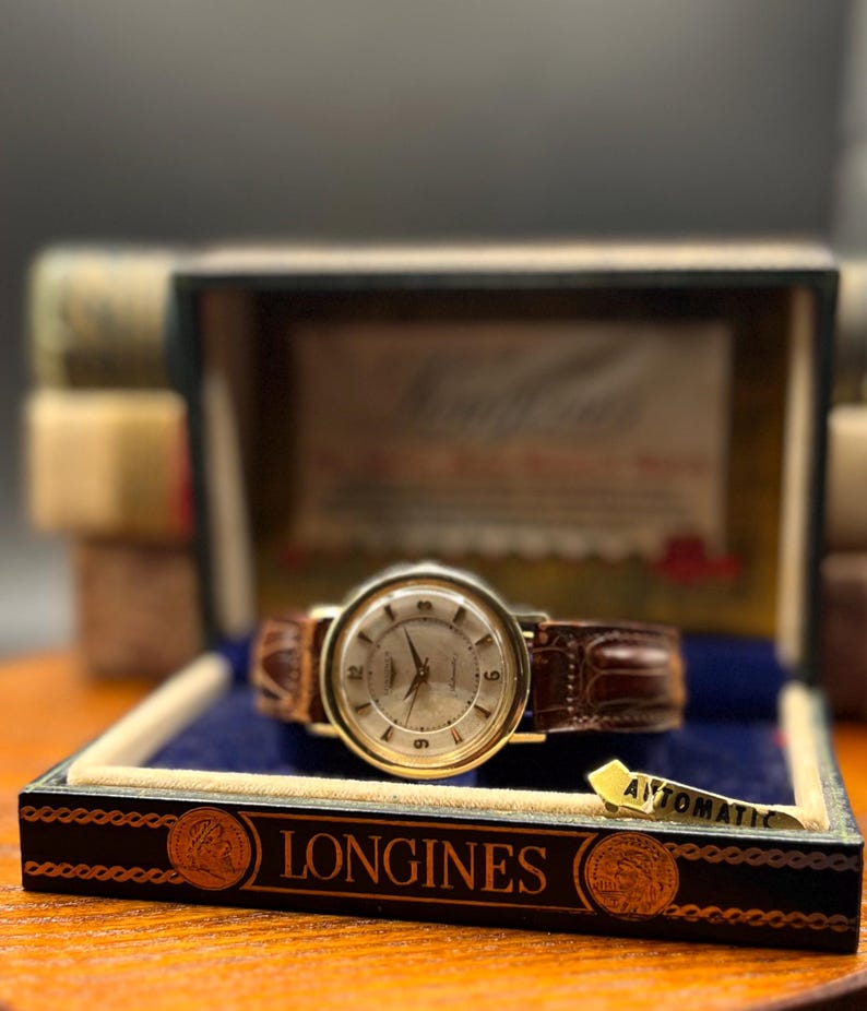 Puede incluir: Un reloj de pulsera autom&aacute;tico Longines vintage con una correa de cuero marr&oacute;n, presentado en su caja original. El reloj tiene una caja dorada y una esfera clara con marcadores de hora. La caja es negra con letras doradas y un interior azul.