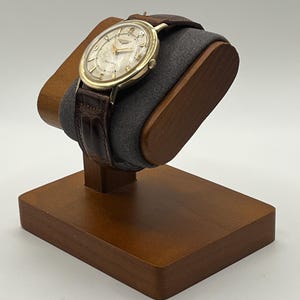 Puede incluir: Un reloj de pulsera dorado con una correa de cuero marr&oacute;n descansa sobre un soporte de madera para relojes. El reloj tiene una esfera de color claro con manecillas y marcadores dorados. El soporte es de madera marr&oacute;n oscuro y tiene un coj&iacute;n gris.