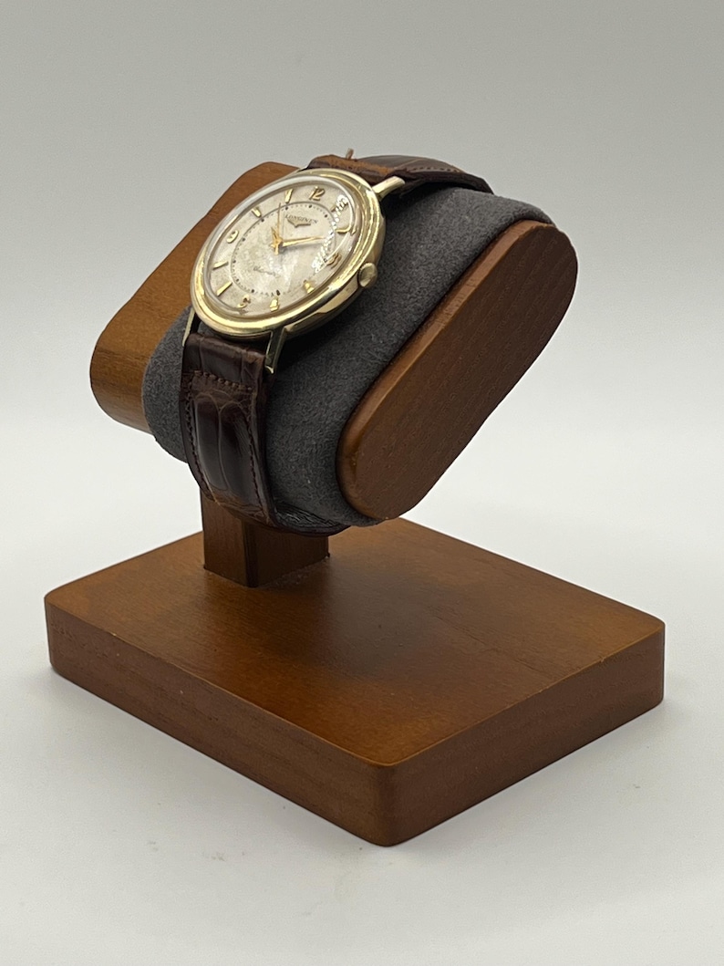 Puede incluir: Un reloj de pulsera dorado con una correa de cuero marr&oacute;n descansa sobre un soporte de madera para relojes. El reloj tiene una esfera de color claro con manecillas y marcadores dorados. El soporte es de madera marr&oacute;n oscuro y tiene un coj&iacute;n gris.