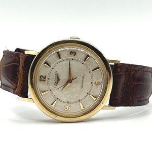 Puede incluir: Un reloj de pulsera autom&aacute;tico Longines vintage en tono dorado con una correa de cuero marr&oacute;n. La esfera del reloj es de color claro con agujas y marcadores dorados. El reloj tiene las palabras "Longines" y "Automatic" en la esfera.