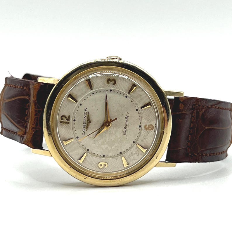 Puede incluir: Un reloj de pulsera autom&aacute;tico Longines vintage en tono dorado con una correa de cuero marr&oacute;n. La esfera del reloj es de color claro con agujas y marcadores dorados. El reloj tiene las palabras "Longines" y "Automatic" en la esfera.