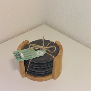 Peut inclure: Ensemble de six sous-verres en ardoise noire avec un support en bambou. Les sous-verres sont liés ensemble avec de la ficelle et portent une étiquette verte et blanche qui indique "Sous-verres en ardoise Base en bambou".