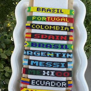 May include: A collection of ten rectangular beaded bracelets arranged vertically on a light-colored display board. Each bracelet features a unique design with various colors and text including BRASIL, PORTUGAL, COLOMBIA, SPAIN, ARGENTIN, MESSI, MEXICO, and ECUADOR.