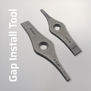 Op de afbeelding: Twee grijze speling installatiegereedschappen met een getextureerd oppervlak. Elke tool heeft een cirkelvormig gat en afmetingen van 6 mm en 15 mm. De tekst "Gap Install Tool" staat verticaal links.