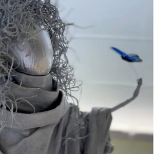 Peut inclure: Une figure sculptée grise avec une surface texturée ressemblant à des cheveux et un long bras tendu. Un petit oiseau bleu s'envole de la figure.