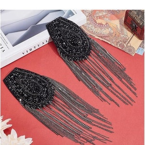 Pode incluir: Dois broches pretos de strass com correntes prateadas penduradas. Cada broche tem formato oval e apresenta um design circular de strass.