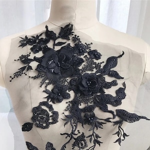 Könnte beinhalten: Schwarze florale Spitzenapplikation mit Perlenbesatz. Die Applikation zeigt eine große Blume, die von kleineren Blumen und Blättern umgeben ist. Die Spitze ist zart und filigran, und die Perlen verleihen ihr einen Hauch von Eleganz.