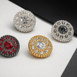 May include: Four decorative buttons with jewel-like embellishments. The buttons feature a variety of colors, including silver, gold, and red. Each button has a central stone surrounded by smaller stones, creating a sparkling effect. The buttons are round and have a classic design.