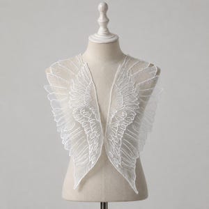 Puede incluir: Una aplicación de encaje bordado en color blanco que presenta un par de alas de ángel simétricas con costuras detalladas de plumas. El diseño de tela fina y transparente se muestra sobre un maniquí, destacando el intrincado trabajo.