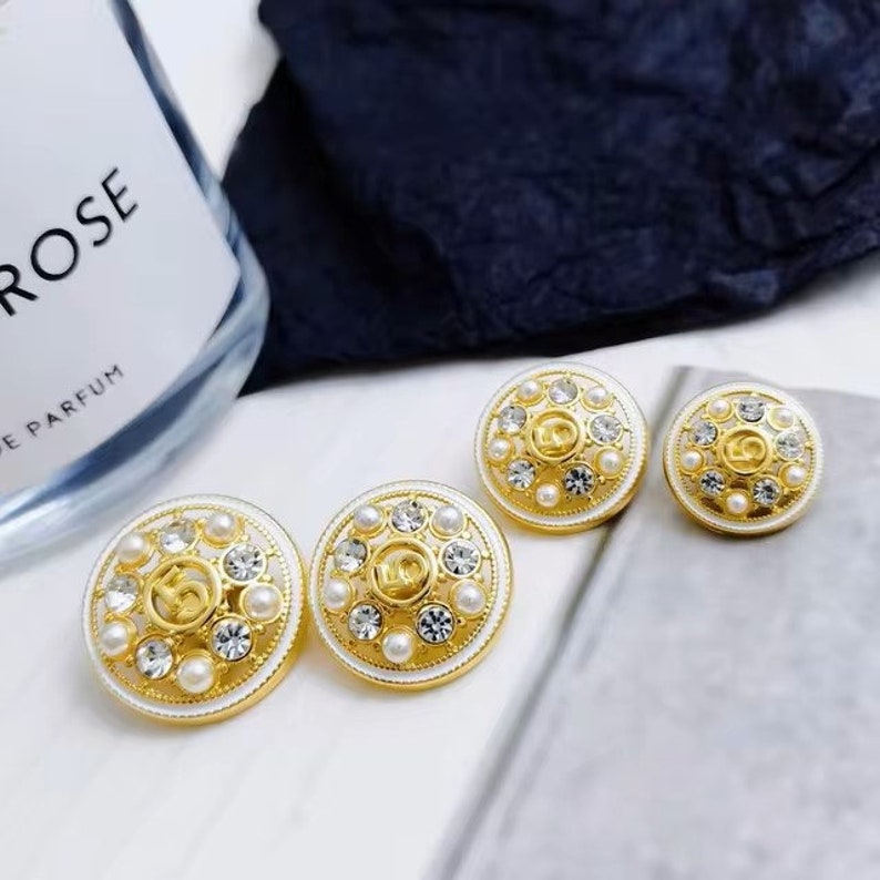 Puede incluir: Tres pendientes de tono dorado con esmalte blanco y detalles de perlas. Los pendientes tienen un dise&ntilde;o circular con un logotipo en relieve en el centro.
