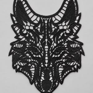 May include: A black lace appliqué shaped like a stylised wolf head. The design features intricate, symmetrical embroidery with openwork patterns forming the ears, eyes, snout, and facial features, creating a detailed, cutout silhouette.
