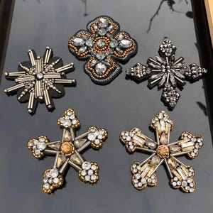 Puede incluir: Un surtido de cinco adornos decorativos con diseños de cruces y estrellas. Presentan cristales plateados, dorados y transparentes, cuentas y pedrería. Cada pieza es única, con formas y tamaños variados.