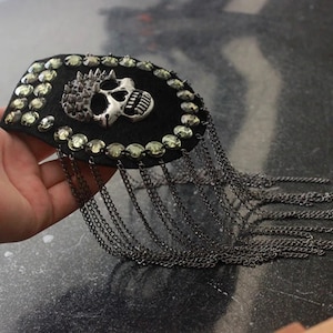 May include: A black skull patch with silver accents and rhinestones, surrounded by a fringe of silver chains.