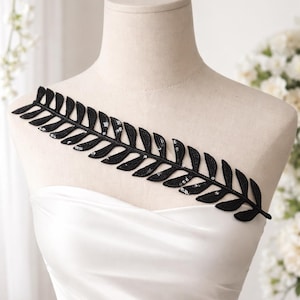 May include: A black sequined leaf-shaped applique, possibly for embellishing garments, is displayed on a mannequin. The applique is made of black fabric and features small, shiny sequins. The mannequin is wearing a white garment.