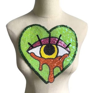 May include: A green heart-shaped sequin patch with a large eye design. The eye is white with a black pupil and yellow iris. The eye is dripping with orange sequins.