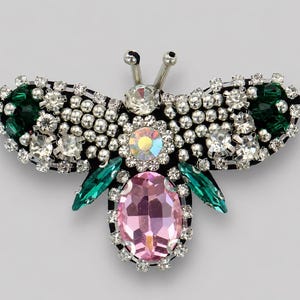 May include: A bee-shaped brooch featuring a large oval pink central crystal, emerald green marquise-cut side stones, and clear round rhinestones. The wings are embellished with silver-toned beads and clear crystals, with two silver antennae on top.