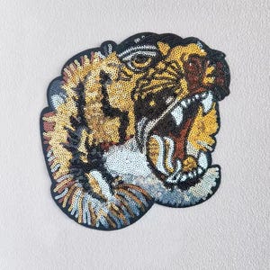 May include: A large, embroidered tiger head patch with a fierce expression. The patch is made of sequins in shades of gold, black, white, and brown.