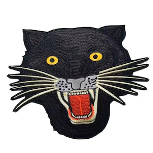 Puede incluir: Parche bordado negro que representa la cara de una pantera con una expresión feroz, ojos amarillos y lengua roja. El parche tiene un fondo blanco y es perfecto para agregar un toque de salvajismo a la ropa o los accesorios.