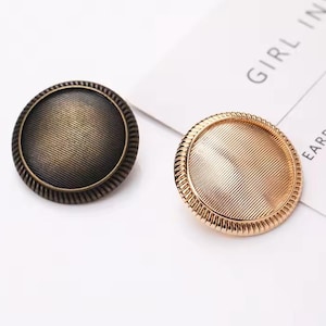 May include: Two round metal buttons with a textured surface. The button on the left is a dark bronze colour. The button on the right is gold.