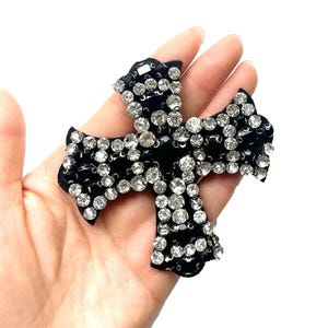 May include: A black cross-shaped patch with rhinestones. The cross is made of black fabric and is covered in clear rhinestones.