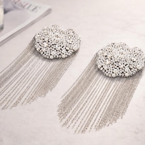 May include: Two cloud-shaped brooches with a cluster of small, shiny, clear beads and dangling silver chains. The brooches are on a light-colored surface, with a lit candle and a magazine in the background.