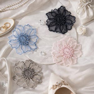 May include: Four delicate, embroidered flower appliques in shades of blue, black, pink, and gray. The floral designs are placed on a white surface with other wedding-themed accessories, including a pair of gold rings, a pearl necklace, and a bridal shoe.