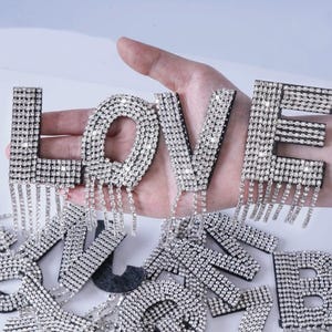 May include: A set of rhinestone-covered letters spelling out "LOVE" with dangling rhinestone chains. The letters are on a hand and there are other letters in the background.