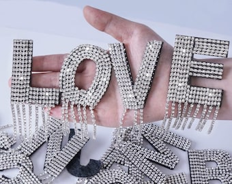 Può includere: Un set di lettere ricoperte di strass che compongono la parola "LOVE" con catene di strass pendenti. Le lettere sono su una mano e sullo sfondo ci sono altre lettere.