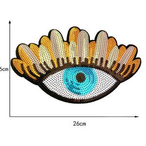 May include: A decorative eye-shaped patch, 16cm high and 26cm wide, with a blue iris and a black outline. The eyelashes are a mix of gold, white, and brown sequins. The eye is embellished with sequins.