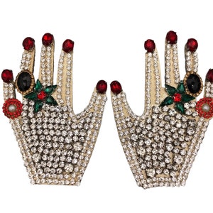 Puede incluir: Un par de broches en forma de mano de color dorado con pedrería, joyas rojas y verdes y detalles en blanco y negro.