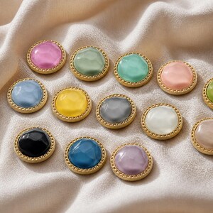 Könnte beinhalten: Eine Sammlung bunter, runder Knöpfe mit einem goldfarbenen, strukturierten Rand. Die Knöpfe weisen eine Vielzahl von Pastellfarben auf, darunter Rosa, Blau, Grün und Gelb. Die Knöpfe sind auf einem cremefarbenen Stoff angeordnet.