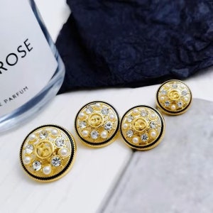 Puede incluir: Tres botones redondos de color dorado con borde negro y detalles de perlas blancas. El centro de cada bot&oacute;n presenta un logotipo dorado con el n&uacute;mero "5" en una fuente estilizada.