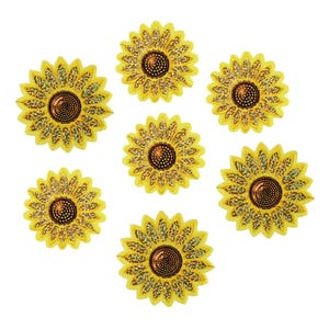 Puede incluir: Siete adornos de girasol amarillos con detalles de purpurina. Cada flor tiene un centro marrón oscuro y pétalos amarillos. Los adornos están dispuestos sobre un fondo blanco.