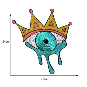 May include: A decorative patch featuring a stylized eye with a gold crown. The eye is turquoise with a black pupil, surrounded by a white dotted pattern. The crown is gold with pink accents. The patch measures 24cm high and 27cm wide.