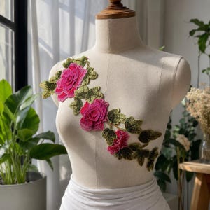 Puede incluir: Aplicación floral decorativa con rosas rosas vibrantes y hojas verdes. La aplicación se muestra en un maniquí, mostrando su diseño y uso potencial en proyectos de moda o artesanía. Las rosas tienen una apariencia detallada y texturizada.