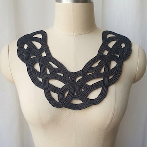 Puede incluir: Un collar negro tejido intrincadamente, exhibido en un maniquí. El collar presenta un diseño de nudo celta, con bucles entrelazados que crean un patrón audaz y texturizado. El color oscuro y la artesanía detallada sugieren una pieza llamativa.