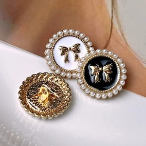 Op de afbeelding: Twee decoratieve knoppen met een parelrand. De ene knop heeft een witte achtergrond met een gouden strik, de andere heeft een zwarte achtergrond met een gouden strik.